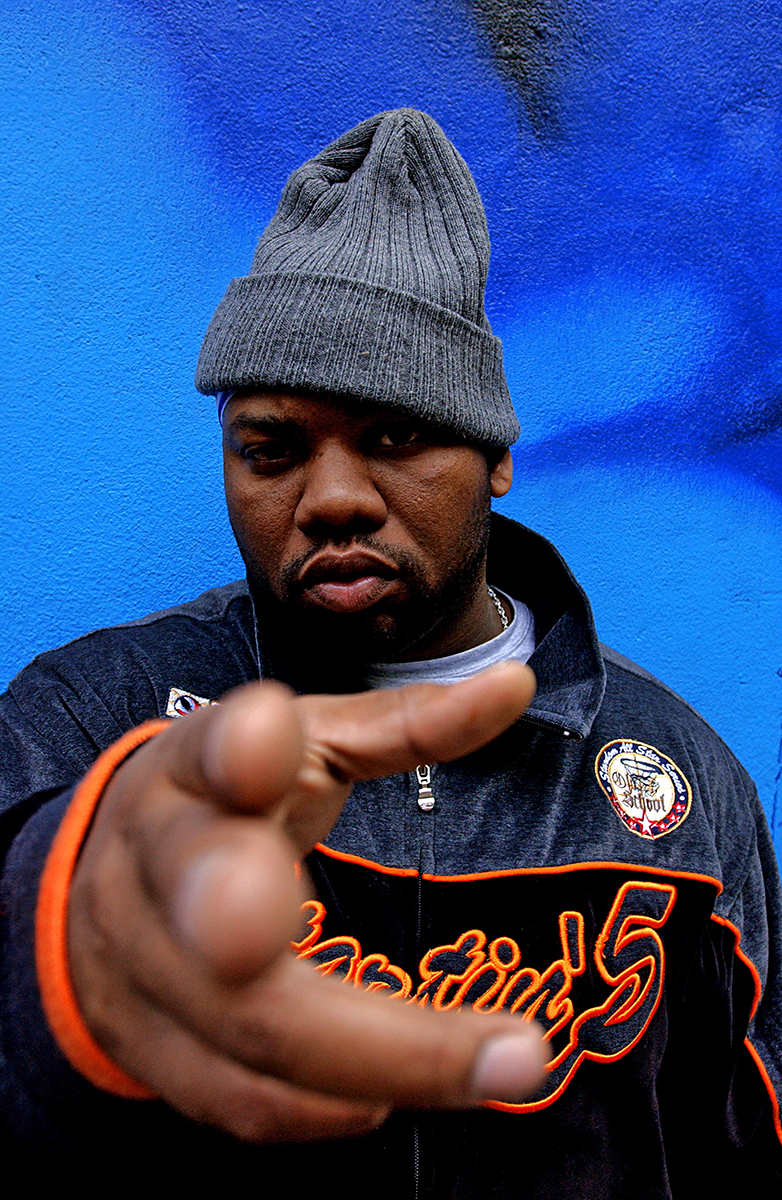 Portrait rapproché de Raekwon du Wu-Tang Clan posant devant un mur bleu, main tendue vers l’objectif