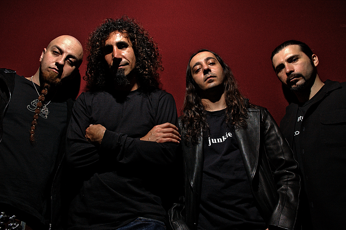 Les quatre membres de System of a Down posent devant un mur rouge, vêtus de noir, dans une ambiance dramatique et sobre.