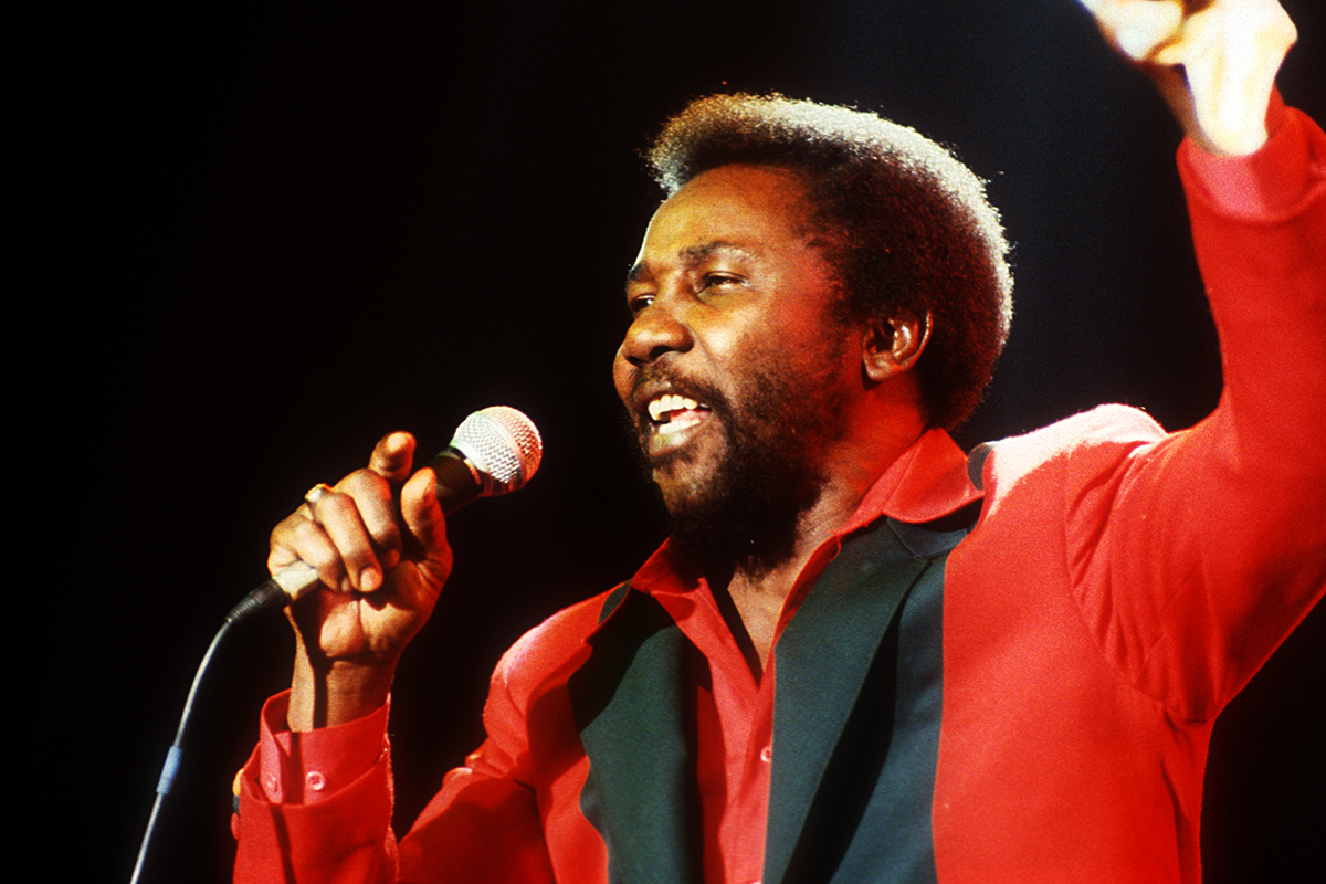 Toots Hibbert de Toots & The Maytals chante en concert, vêtu d'une veste rouge.