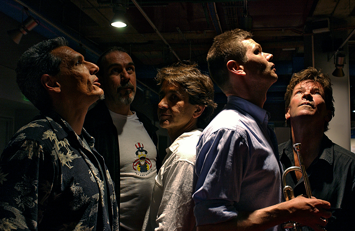 Les membres de Tuxedomoon posent dans une ambiance sombre et théâtrale, éclairés par une lumière venant du plafond, l’un d’eux tenant une trompette.