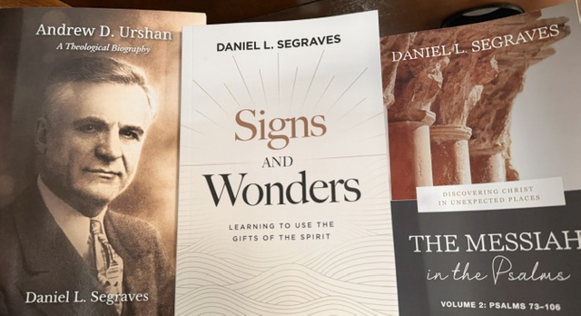 A collection of three books on a wooden surface, including a biography of Andrew D. Urshan, a book titled 'Signs and Wonders' by Daniel L. Segraves, and 'The Messiah in the Psalms, Volume 2' by Daniel L. Segraves.