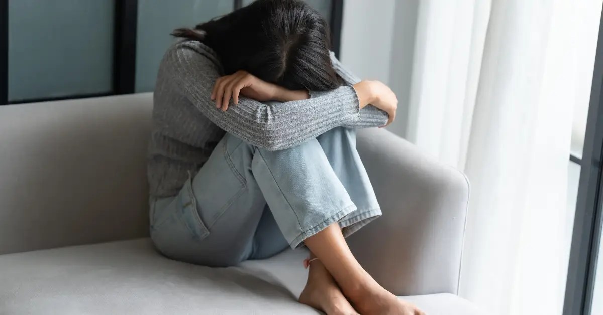 Young woman sitting curled up on a couch with her head on her knees, looking overwhelmed with sadness, representing grief counseling in Enid, OK.