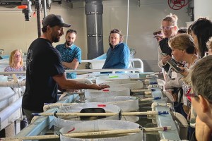 Rutgers AIC Hatchery Manager, Sean Towers, standing in front of a row of vats filled with oysters and water. He holds a handful up to a group of workshop participants crowding around the vats.