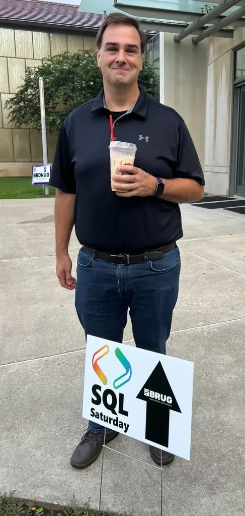 Jason Romans standing in behind the SQL Saturday BRUG sign holding iced cofee