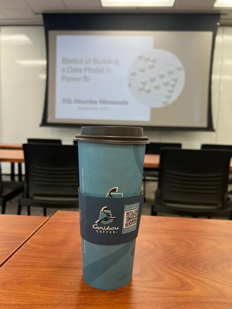 Coffee cup in focus with Caribou Coffee logo in front of a blurry slide titled Basics of Building a Data Model in Power BI