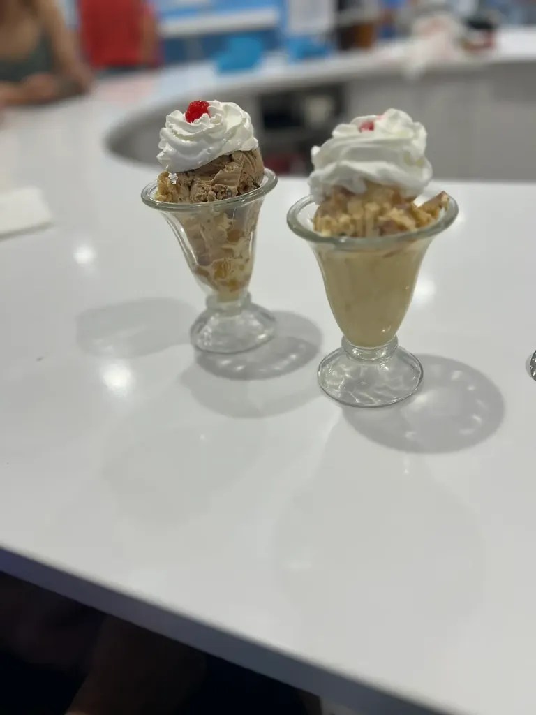 Two ice cream sundaes with whipped cream and toppings on a white table.