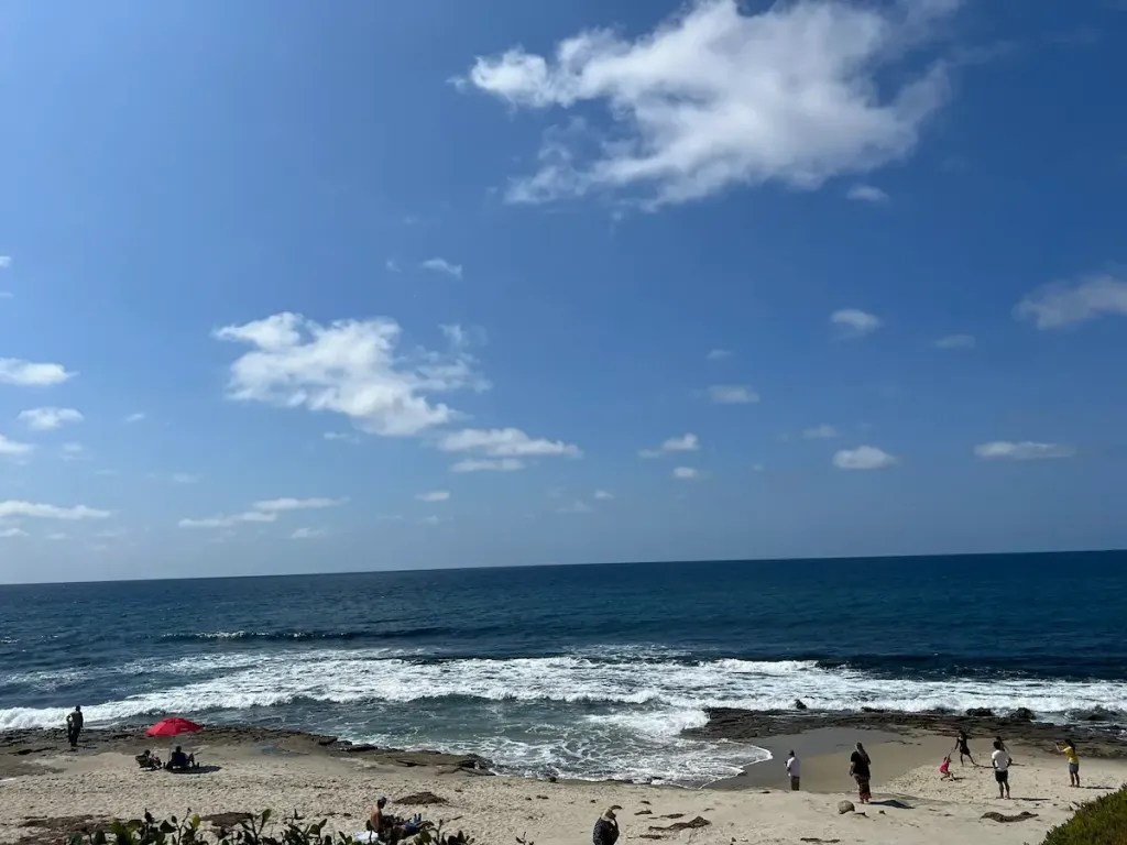 Sand with people playing and looking out at the water - the water is white with small waves and then extends out and is a dark blue