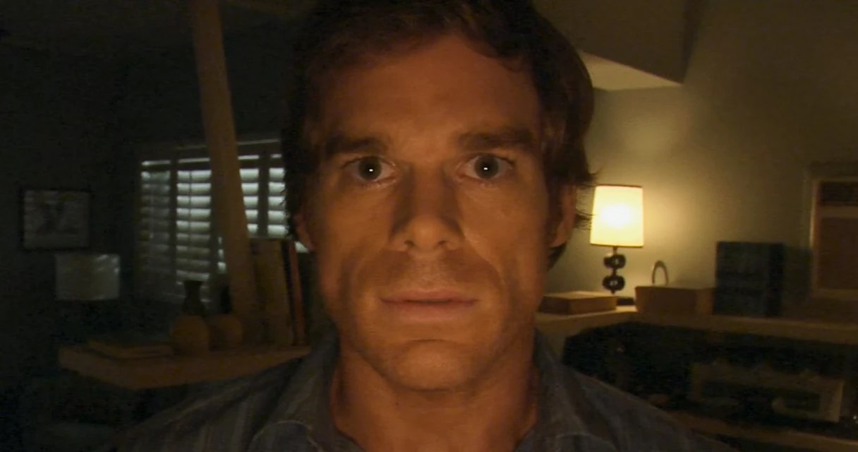 Dexter Morgan from Dexter sitting in a dimly lit room with a lamp behind him, staring blankly into the camera, no text