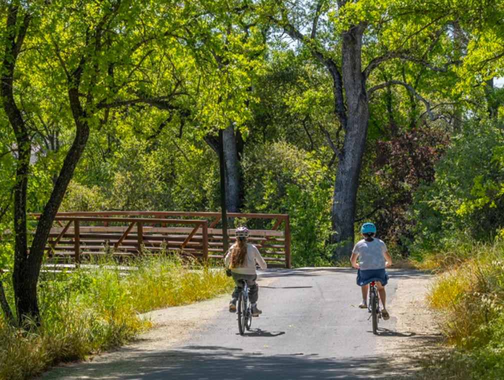 Two bicyclists riding on the Arcade-Cripple Creek Trail in Citrus Heights, California, near a wooden bridge and trees.