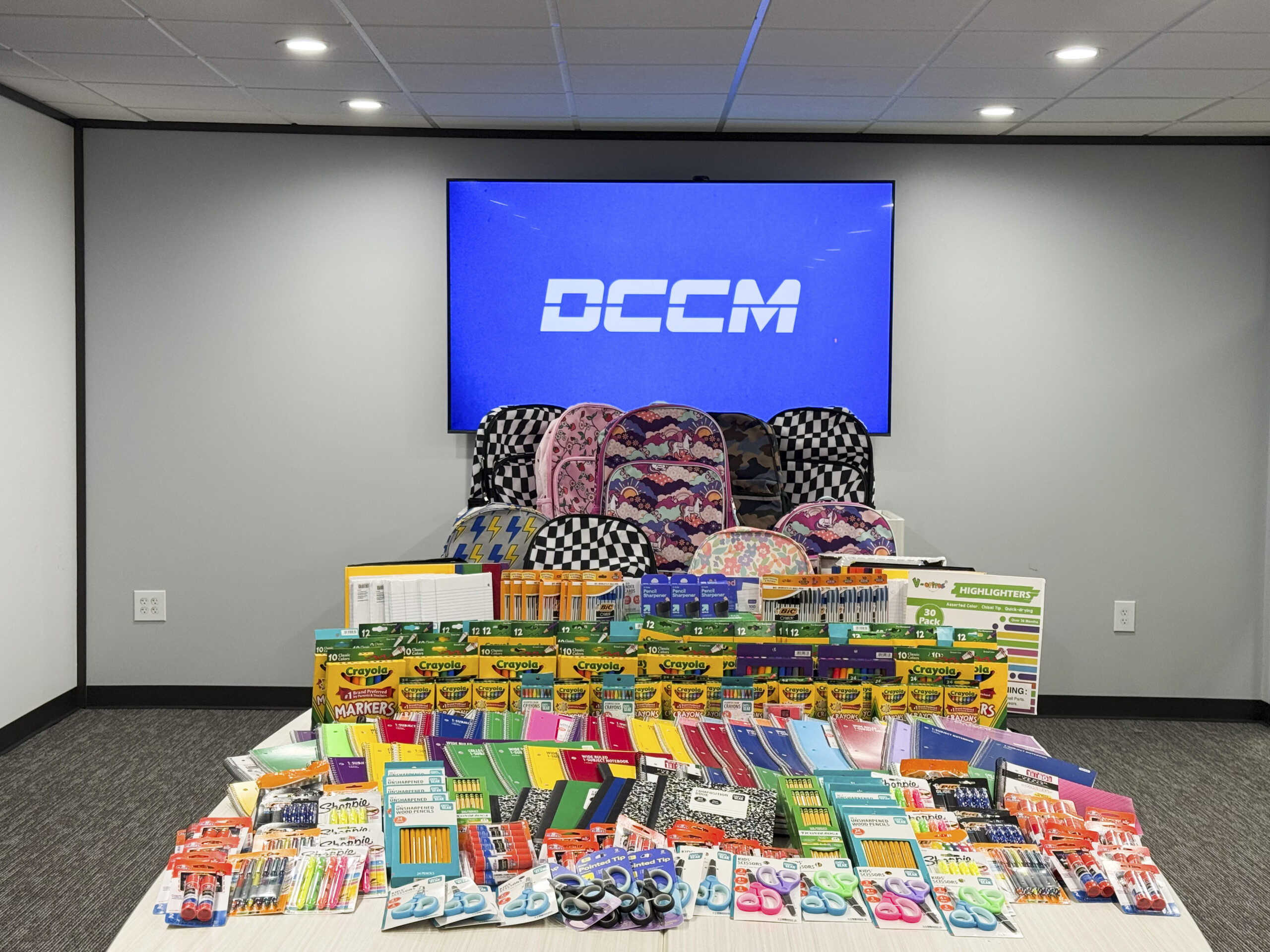 Donated backpacks and school supplies arranged on a table in front of a DCCM screen.