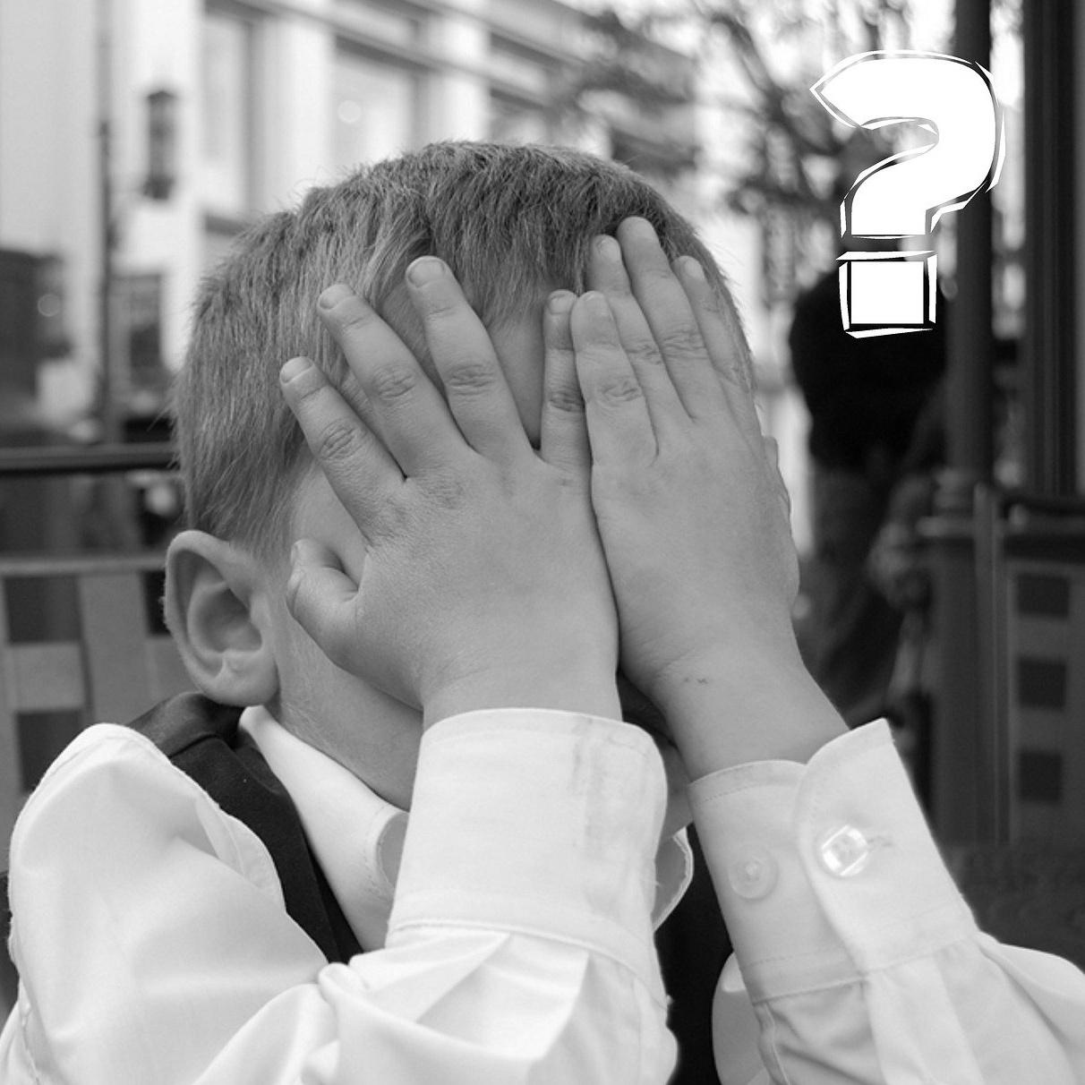 A black and white photo of a well-dressed young boy, burying his face in his hands, with a white question marks to his right