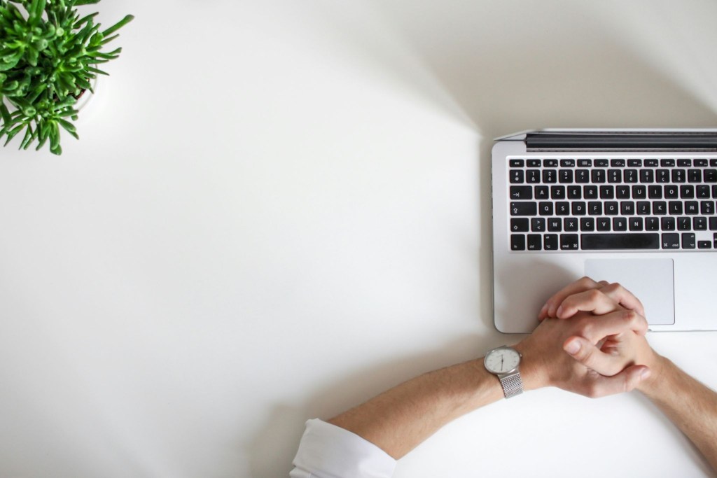Overhead shot of a white laptop on white desk, with a man's hands clasped, resting on the mousepad.