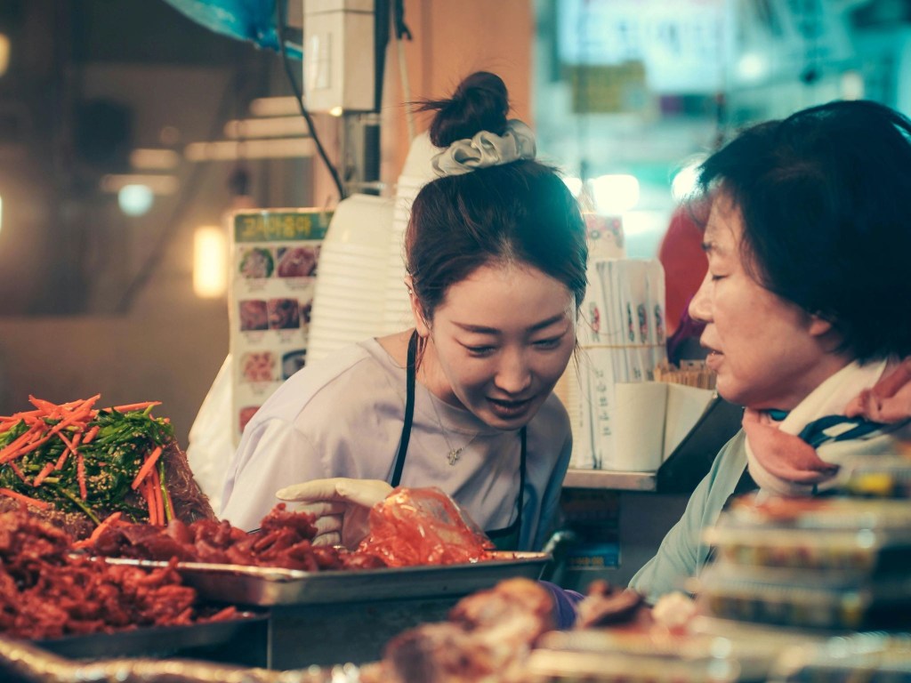 A younger woman and an older woman talking behind trays of kimchi in a restaurant