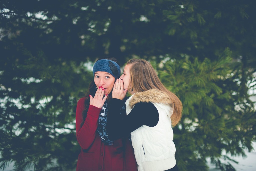 Two women outside, with one whispering in the other's ear, sharing a secret.