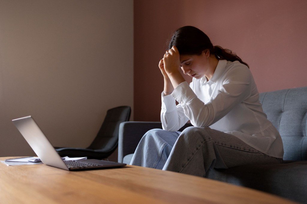 A woman, sitting on a sofa, staring a laptop with with her head in her hands