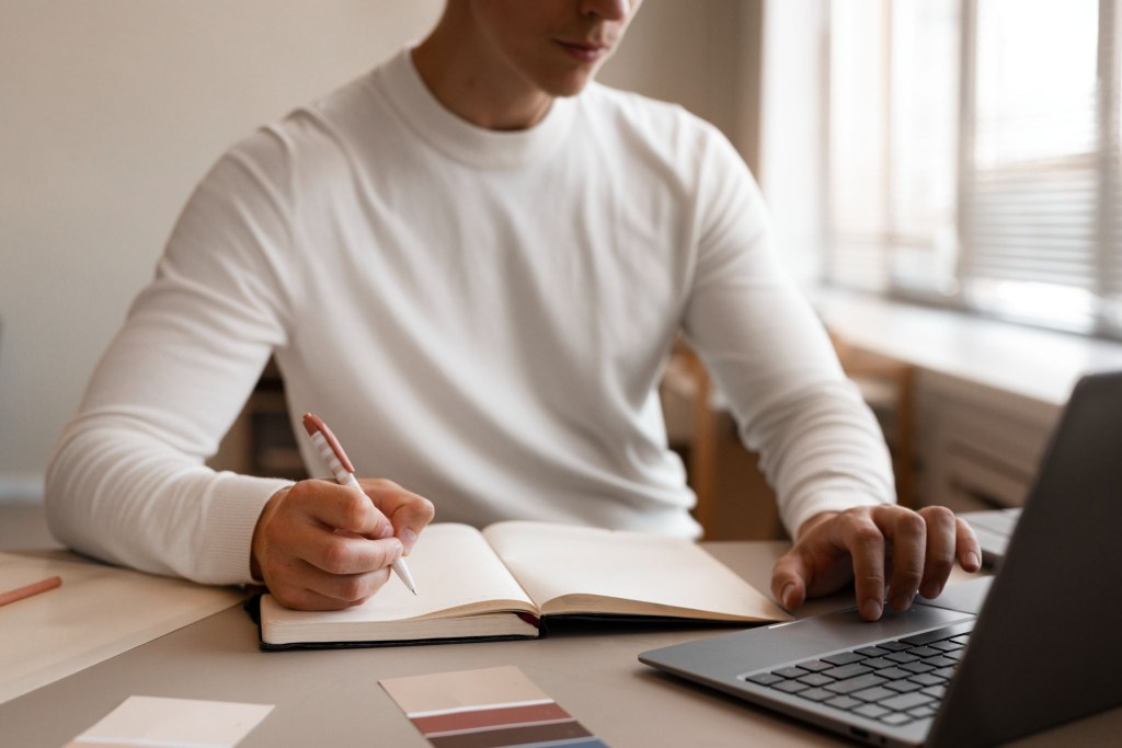 A man sitting at a desk, using a laptop with his left hand and taking notes with his right, perhaps proofreading