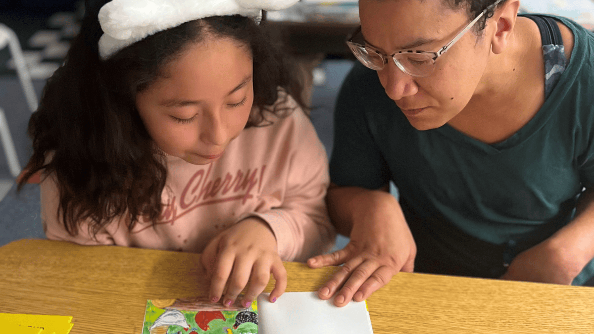 A child wearing bunny ears and a pink sweater with the word "Cherry" on it looks down at a book with a grown-up wearing glasses and a green shirt.