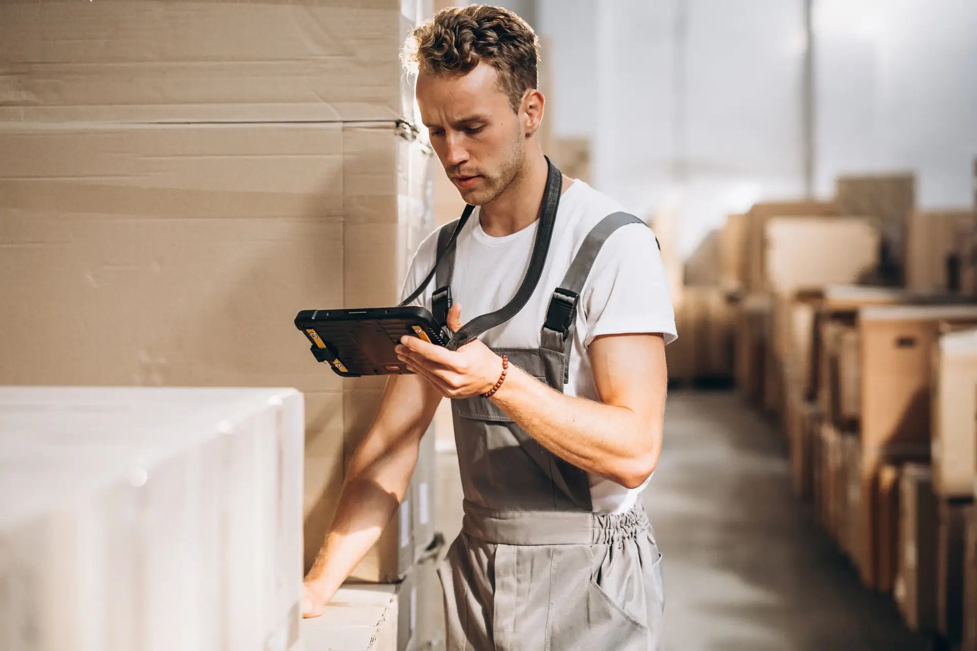 Warehouse worker in gray overalls uses a rugged tablet to check inventory beside stacked cardboard boxes.