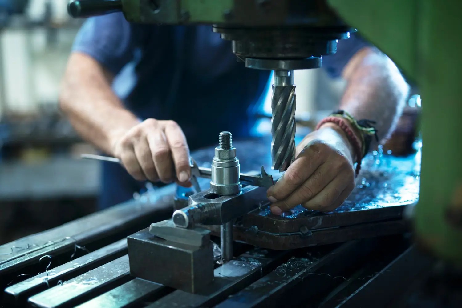 Machinist uses a drill press to bore into a metal part, with calipers on the workpiece nearby.