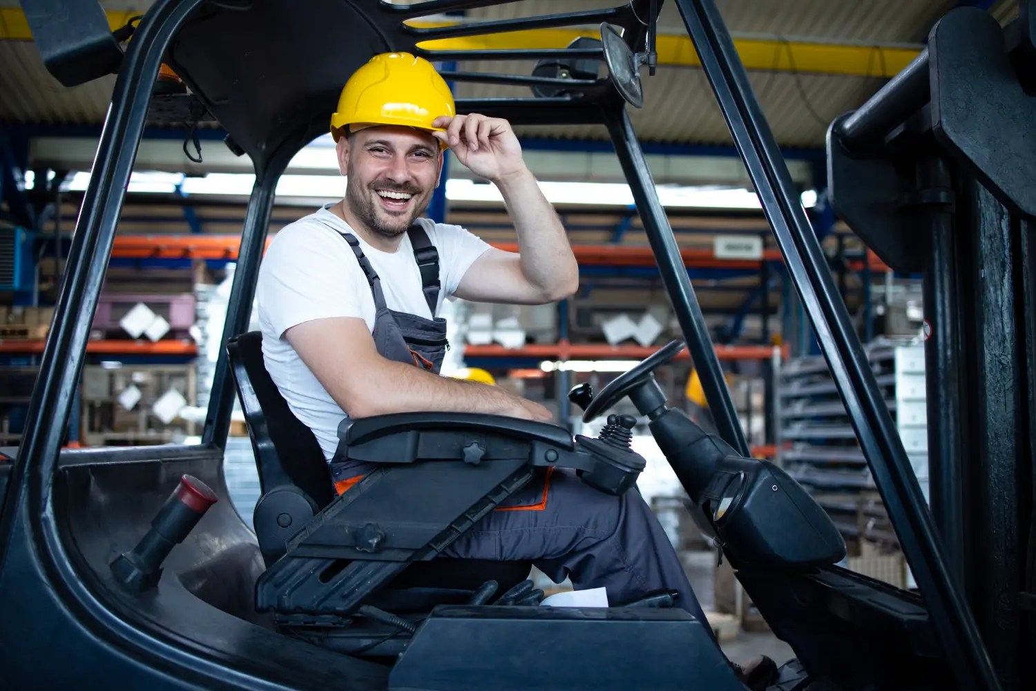 Smiling forklift operator wearing a yellow hard hat sits in the cab inside a busy warehouse