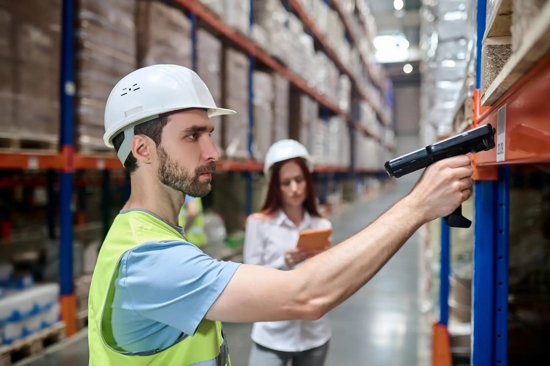Warehouse worker in a white hard hat and high-visibility vest uses a handheld scanner to check inventory on a rack; another person with a clipboard is blurred in the background.