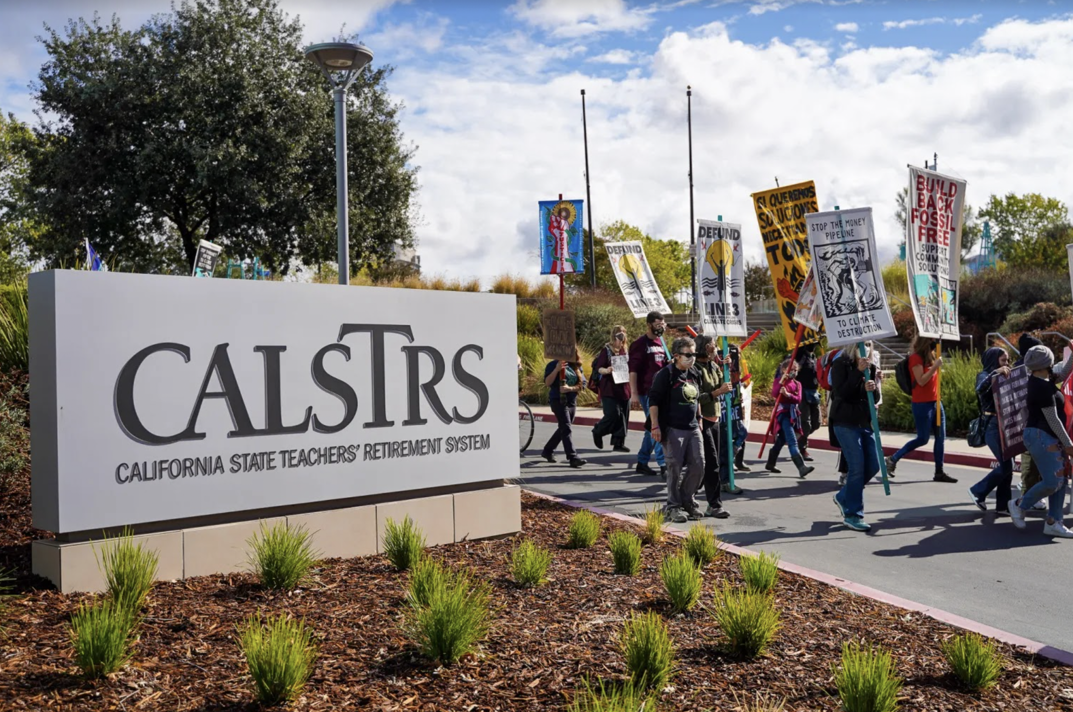 A group of diverse protesters is gathered outdoors, holding signs and banners advocating for fossil fuel divestment.