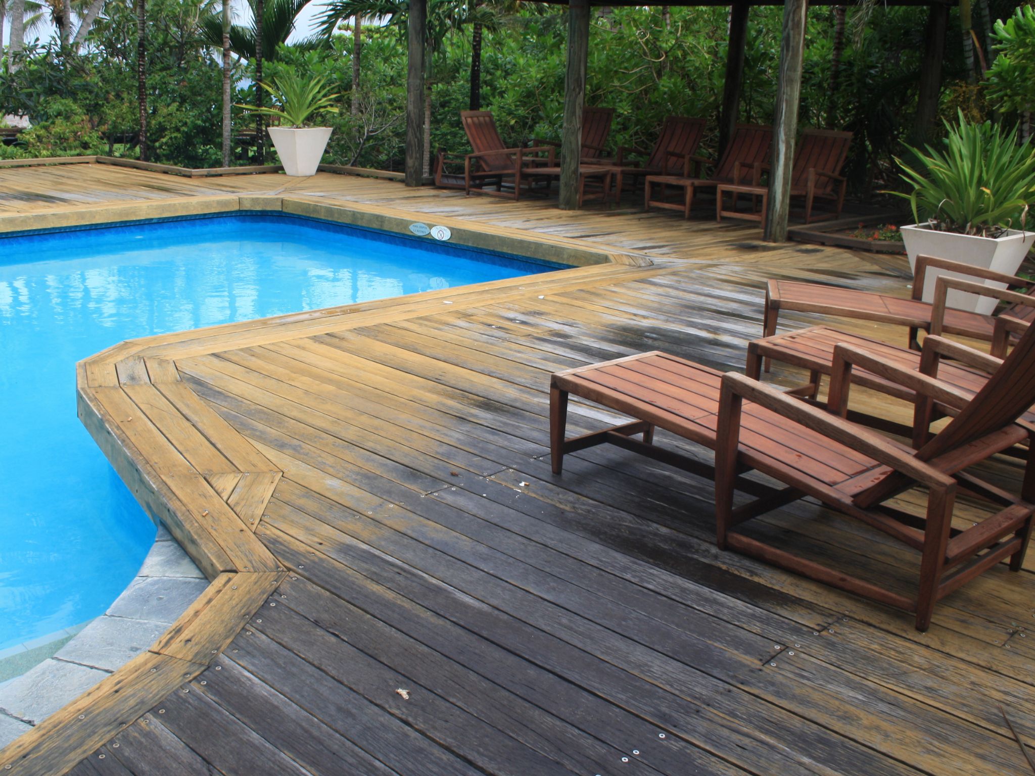 Weathered timber poolside deck with outdoor chairs and lush tropical garden.