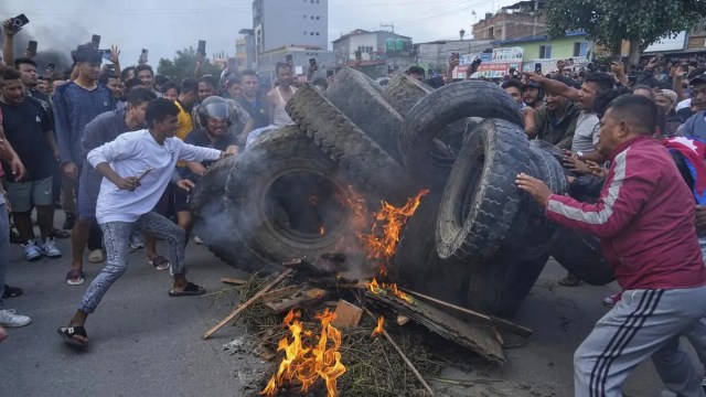 A large crowd of young people, often referred to as "Gen Z," is gathered on a street in Nepal to protest. Many are holding placards with slogans and waving the red and blue national flags of Nepal. The demonstration is a protest against the government's ban on social media and wider issues of corruption and nepotism.