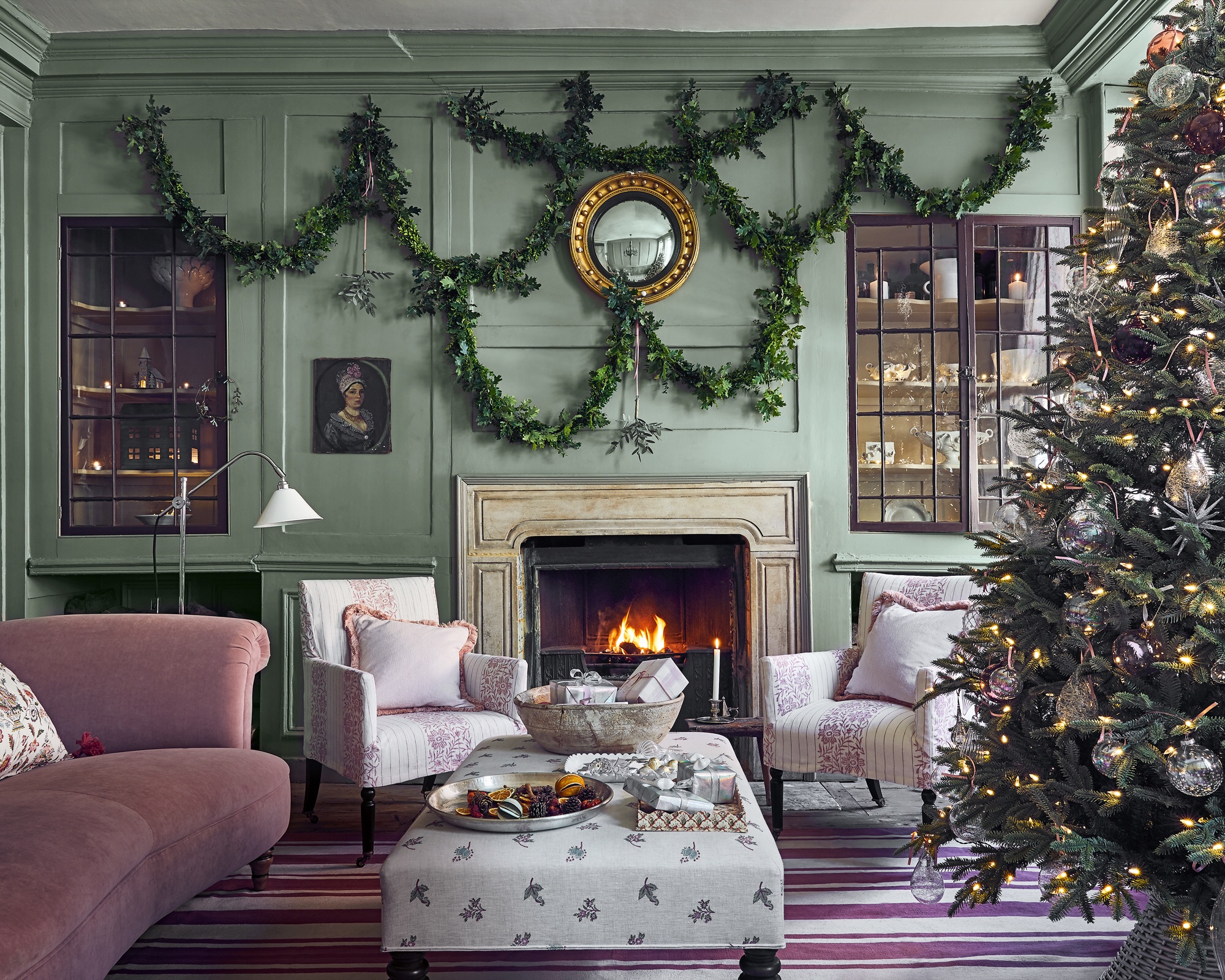 Living room with green walls, green mantelpiece and lit fire with armchairs either side, central ottoman beside decorated Christmas tree.