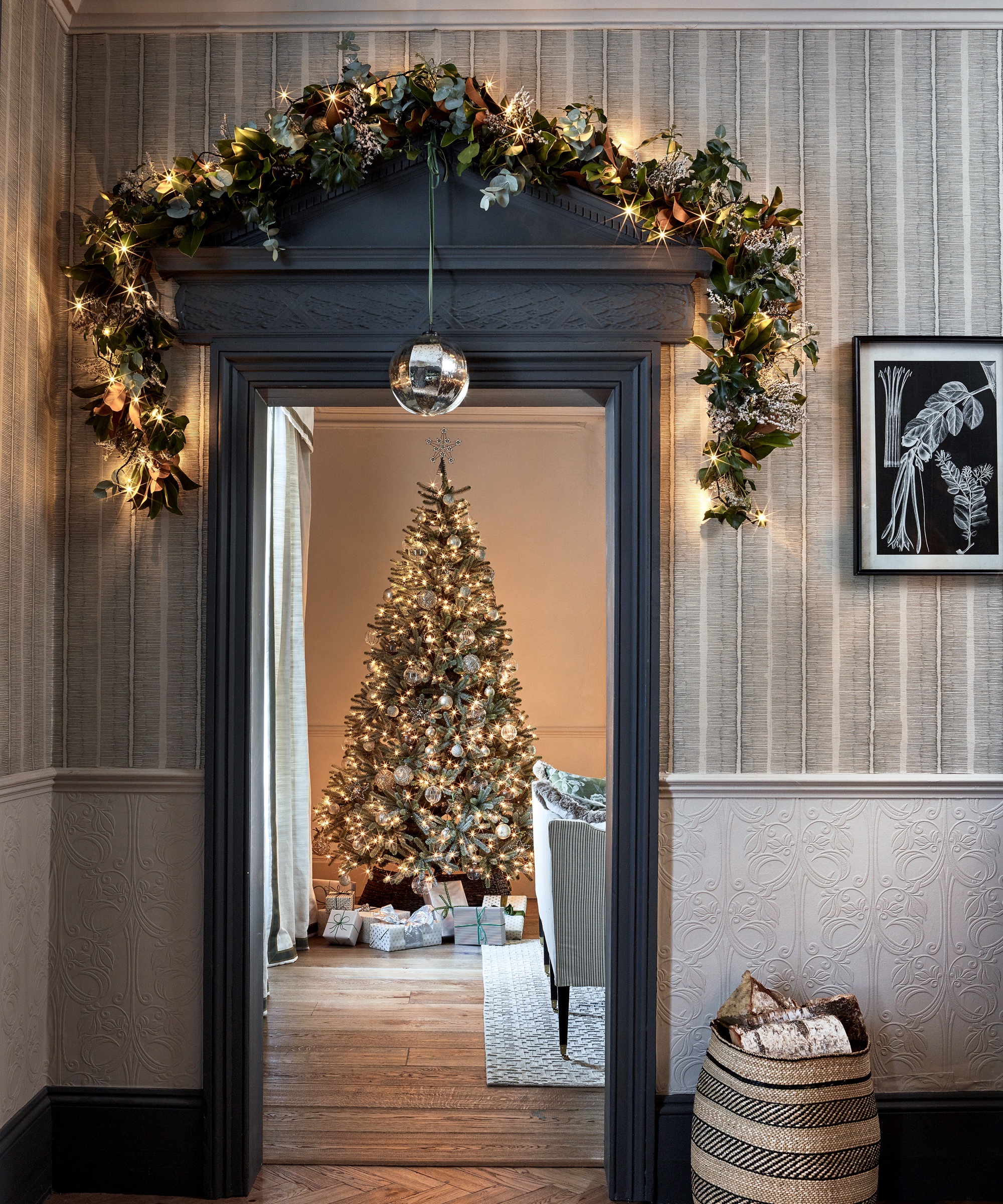 A Christmas doorway with a garland made of greenery threaded with lit golden fairy lights over the lintel, a large crystal bauble hanging down. Pub Orig.