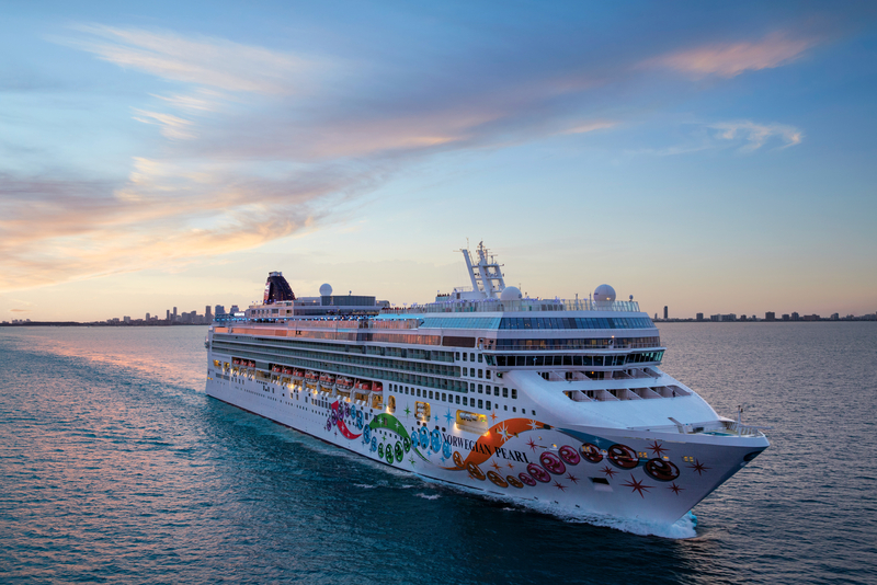 El crucero Norwegian Pearl navegando por el mar durante el atardecer con la silueta de una ciudad al fondo.
