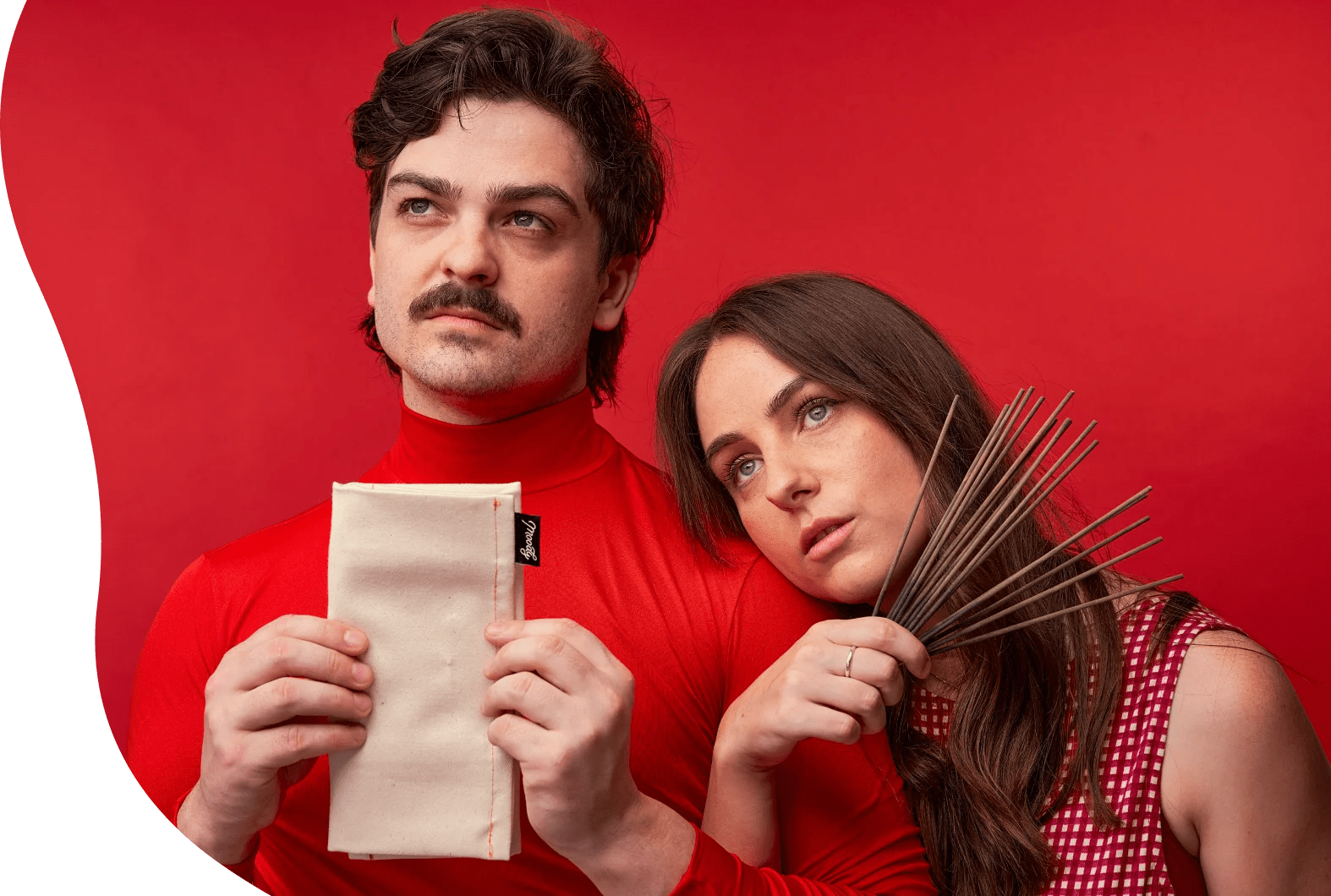 Photo of a man and woman in red turtlenecks against a red background, both with deadpan expressions, the man holding a canvas Moody pouch and the woman holding a fanned bundle of incense sticks