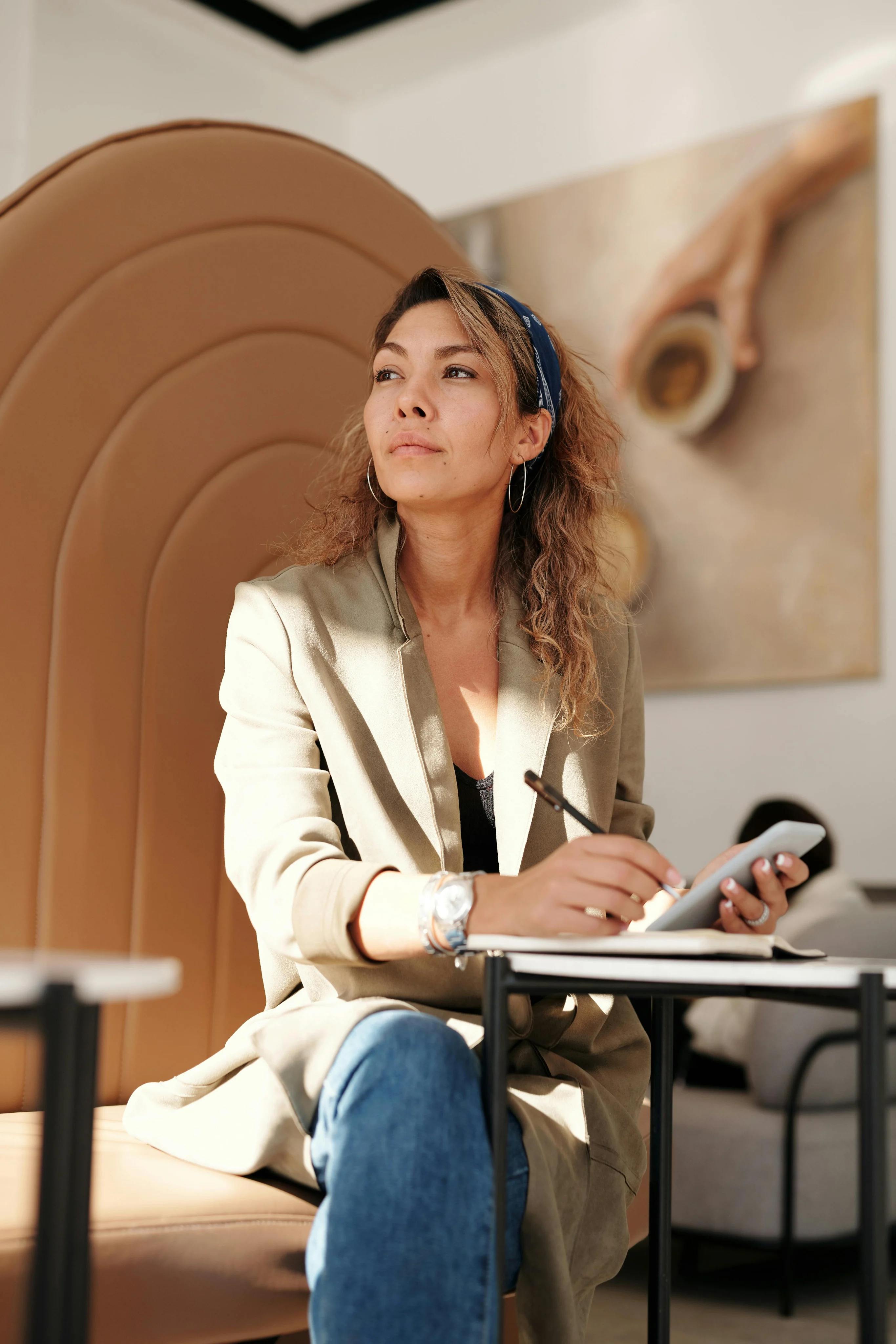 A creative woman at a table, jotting down ideas on her phone and notebook.