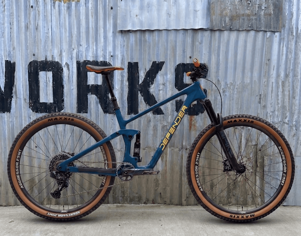 A studio product shot of a teal Defender Orbit full-suspension trail mountain bike with yellow lettering, positioned against a rustic, weathered corrugated metal wall with faint black painted stenciled letters. The bike features matching tan-wall Maxxis Aggressor 29MT tires on both the front and rear wheels, an orange and black saddle, and orange handlebar grips. Below the main bike image, a row of smaller technical component diagrams and a blank metallic badge are displayed on the concrete floor.