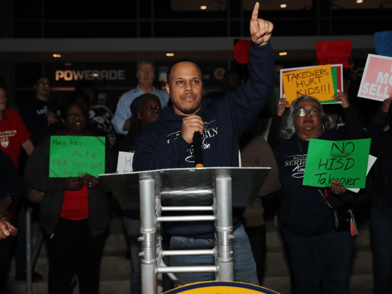 One man stands behind a podium and takes the mic as fellow community members stand behind him in front of Delmar Stadium
