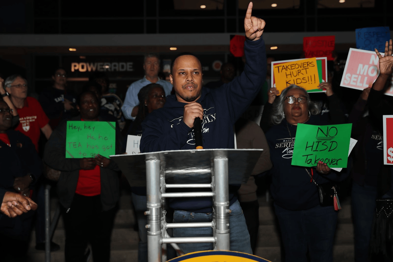 One man stands behind a podium and takes the mic as fellow community members stand behind him in front of Delmar Stadium