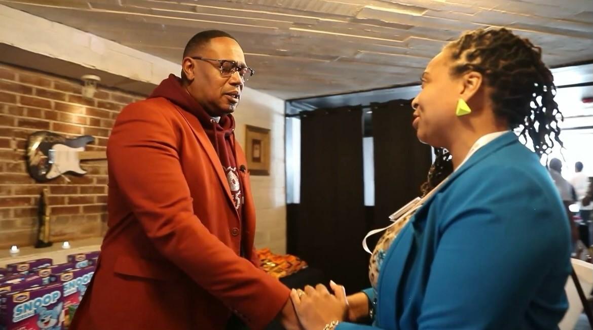Percy "Master P" Miller dressed in a burnt orange suit over a red hoodie top, shakes hands and talks with a woman with locs, green earrings and a turquoise blazer over a blouse