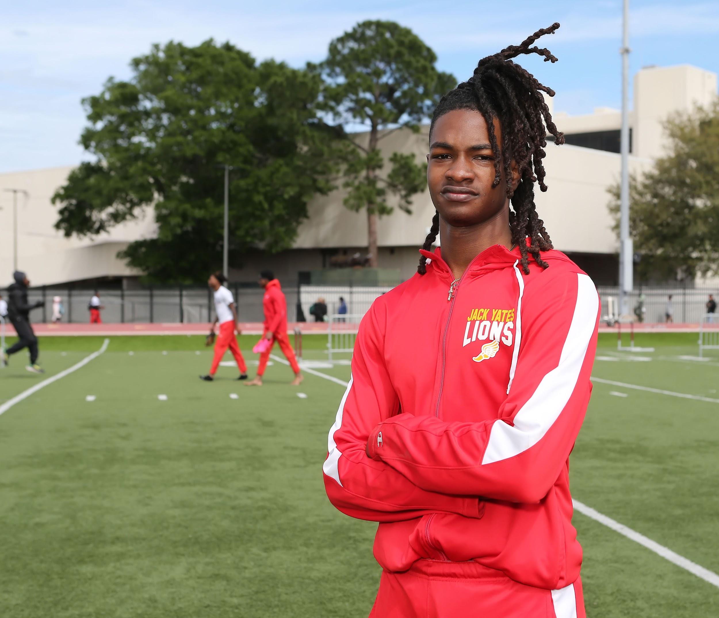 Jeffery LeGrand poses with arms folded on a track field, dressed in a red track suit with a wide white stripe on each sleeve
