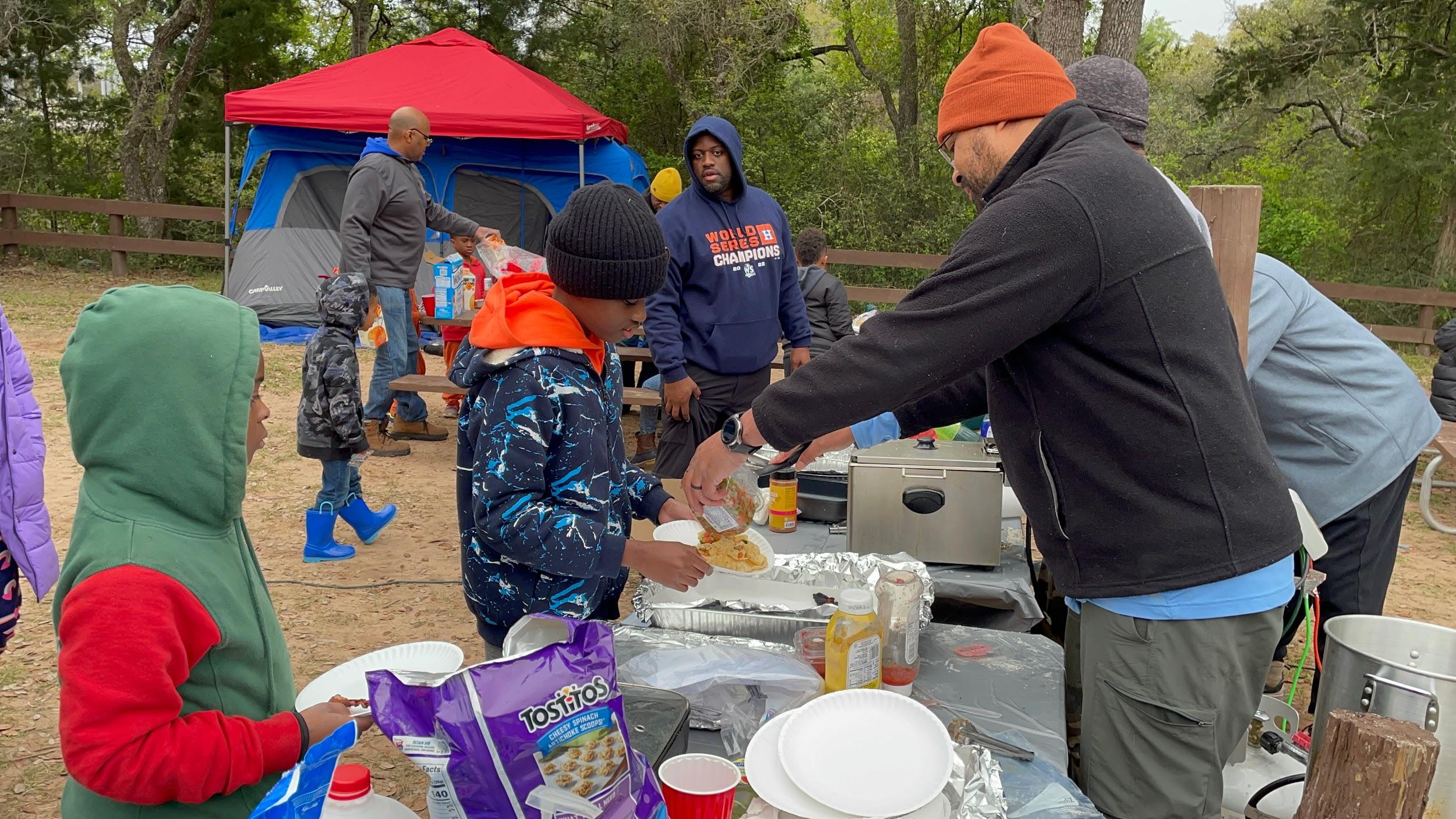 Men serve children food at an outdoors campsite