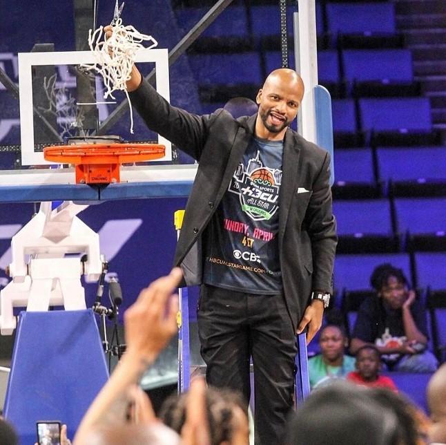HBCU All-Star Game founder Travis Williams holds a shredded basketball net balled up in his hand above the rim of the basketball hoop in gym in front of a crowd during the HBCU All-Star Game.