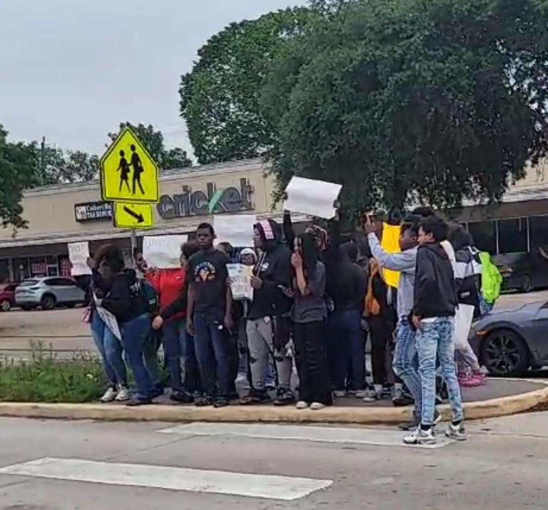 A group of young Black teens stand outside on a street corner in with protest signs