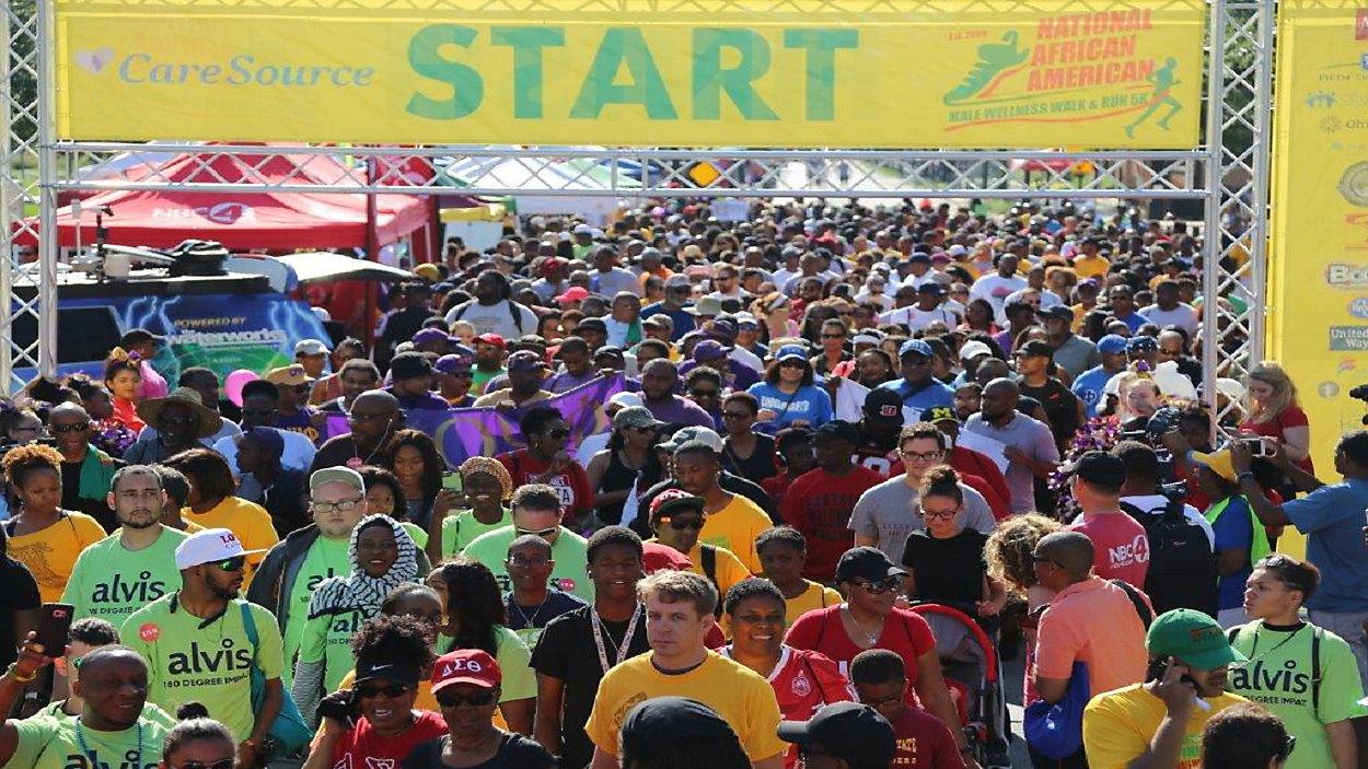 A large crowd walks through the starting point, which is highlighted by a yellow hanging banner with the word "Start"in green font