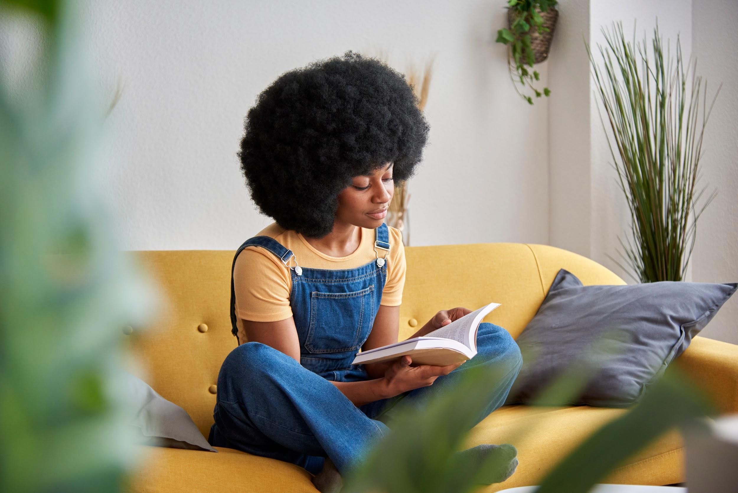 Young black woman sits on a yellow couch in living room reading a book