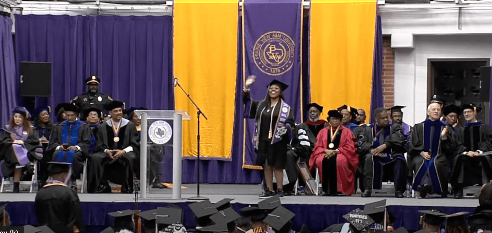 Female college graduate waves to the crowd as she walks across the stage to receive degree