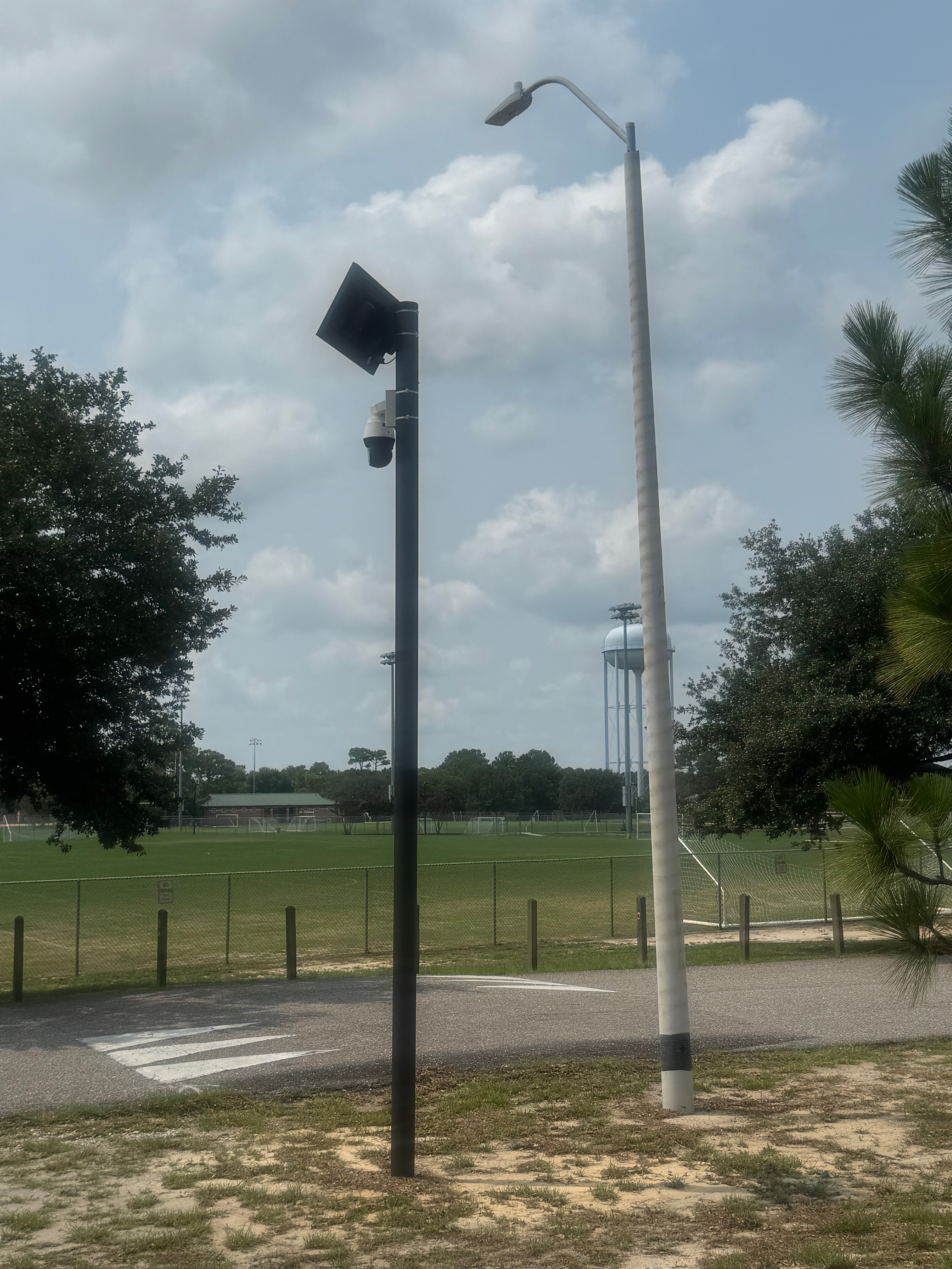 A tall black pole beside a streetlight at Veterans Park; mounted on it are a solar panel, a battery box, and a white pan-tilt-zoom dome camera, with youth soccer fields and a water tower behind.