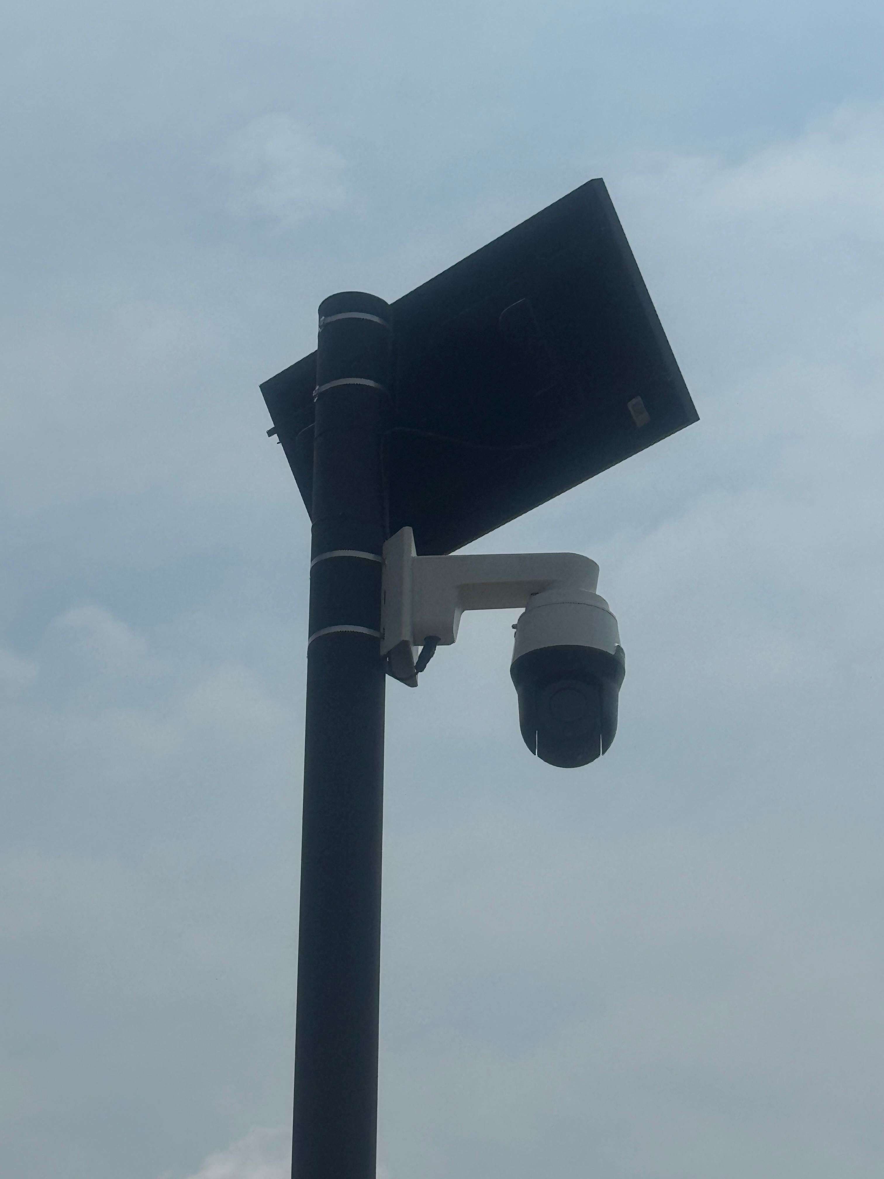 Close-up of the white pan-tilt-zoom dome camera and the solar panel mounted on the black pole against the sky.