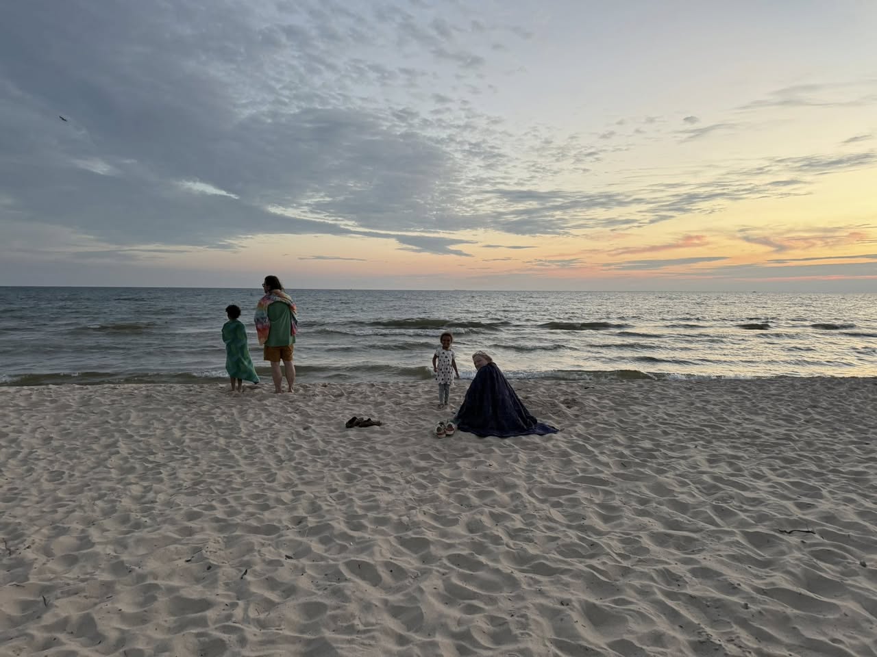 Delight sitting on the beach wrapped in a blue throw style blanked with several people standing around her while a bluish sunset over the lake.