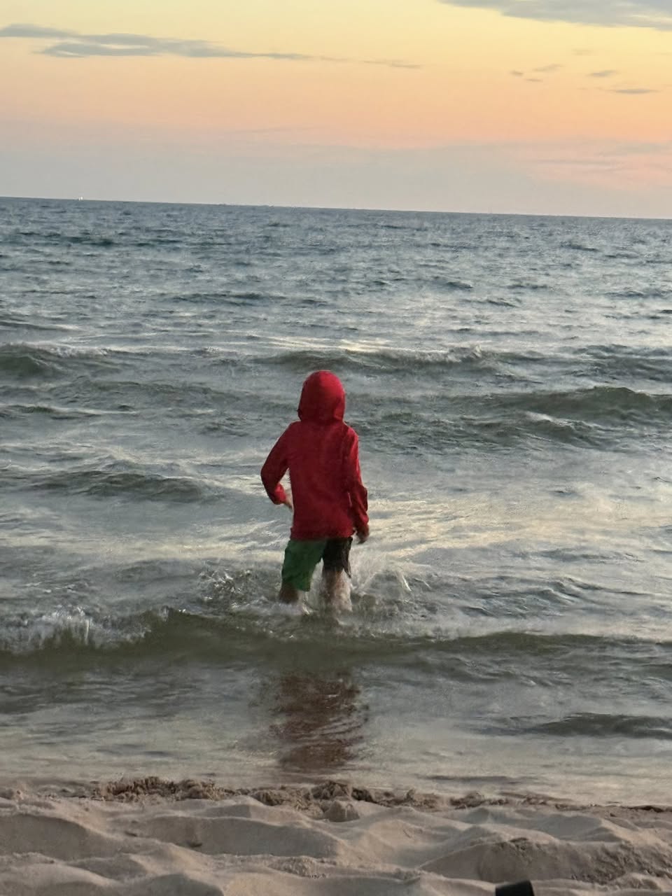 Photo of a human wearing a red colored hooded sweatshirt walking in the lake facing a setting sun.
