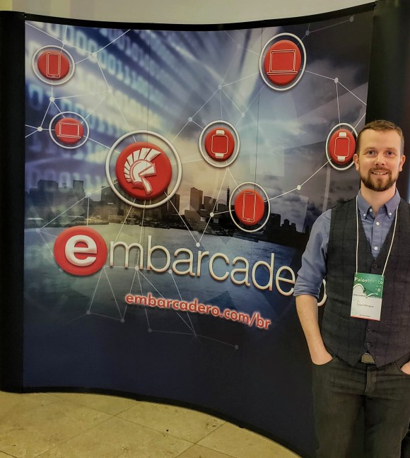 David Millington in front of a sign that says Embarcadero.com/br with a cityscape and reg icons for Delphi and different mobile and desktop platforms