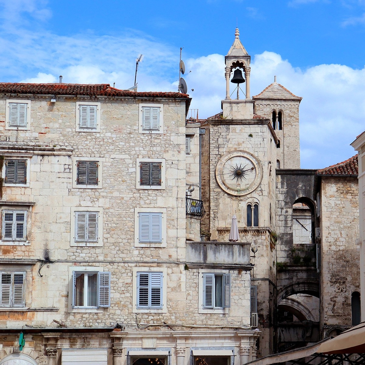 Edifício histórico de pedra com torre do relógio e torre sineira em Split, Croácia. Fachadas desgastadas e venezianas acrescentam um charme rústico sob um céu azul.